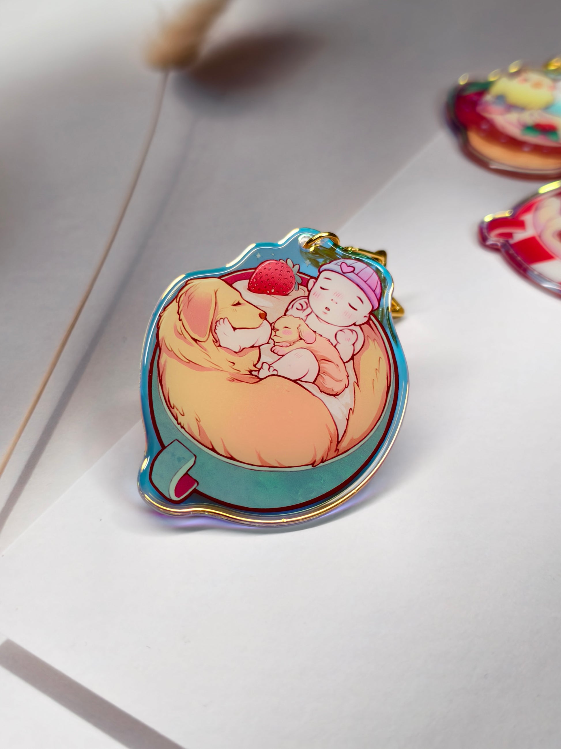 A colorful enamel pin shaped like a teacup, featuring an illustration of a baby sleeping on a yellow blanket with a strawberry, displayed on a white surface.