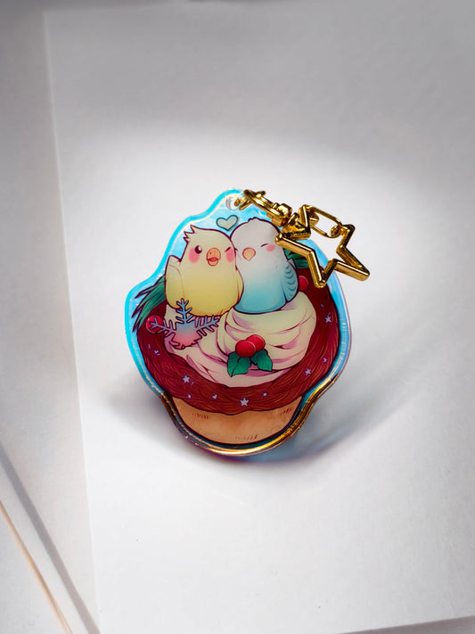 A small enamel pin or charm shaped like a cupcake with two cute pastel birds hugging inside, attached to a gold keychain. The pin is photographed on a white card against a soft white background.