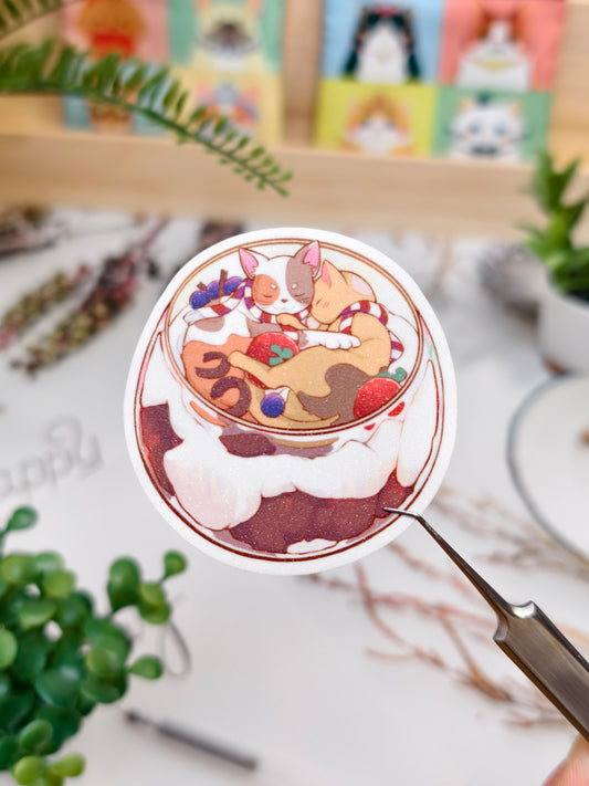 Die-cut vinyl sticker of cute cats inside a layered tiramisu dessert cup held over a wooden desk setup.