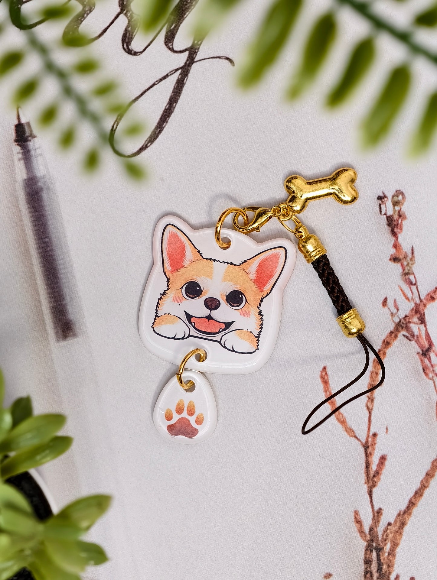 Corgi dog illustrated acrylic phone charm with a dangling paw print charm on a light surface with stationery.