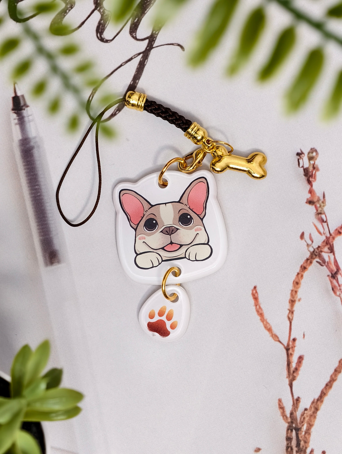 Frenchie dog illustrated acrylic phone charm with a dangling paw print charm on a light surface with stationery.