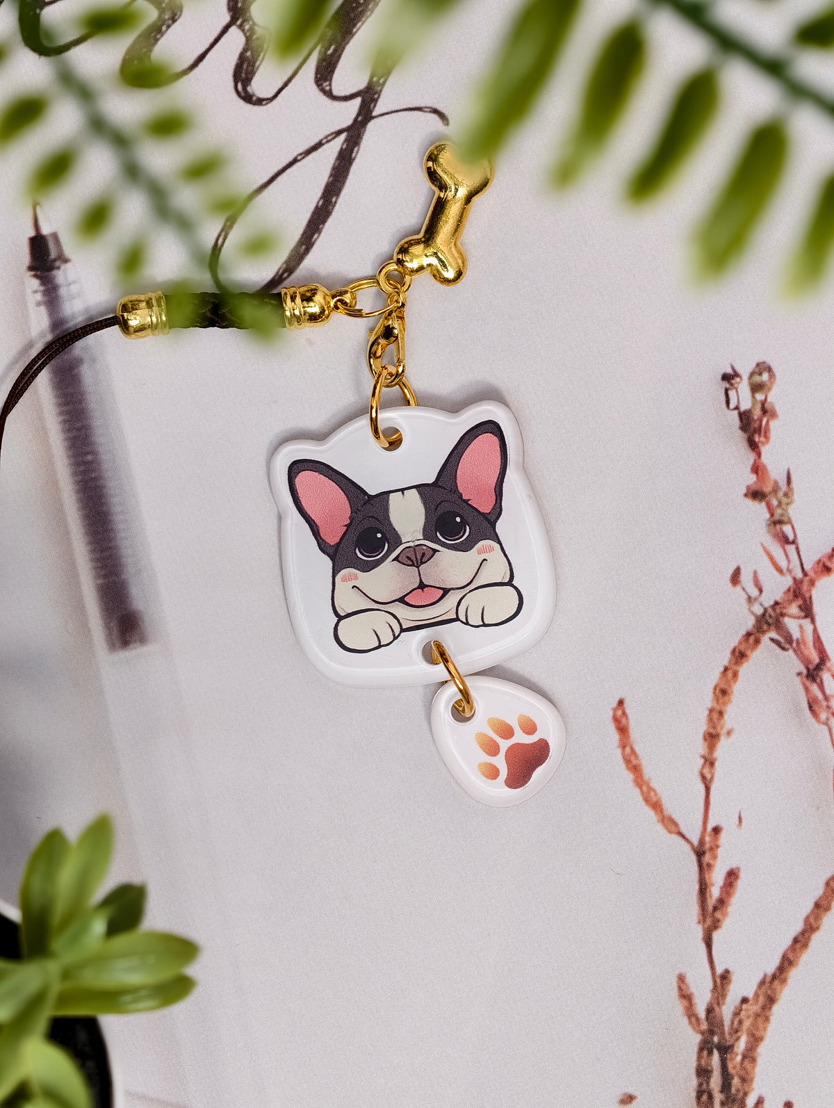 Frenchie dog illustrated acrylic phone charm with a dangling paw print charm on a light surface with stationery.