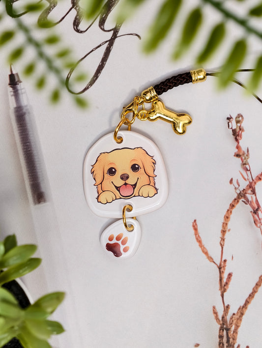 Golden Retriever dog illustrated acrylic phone charm with a dangling paw print charm on a light surface with stationery.