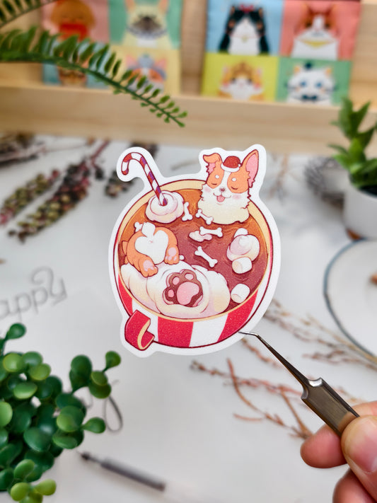 Die-cut vinyl sticker of a cozy Corgi dog relaxing inside a red hot chocolate mug with marshmallows, held over a desk.