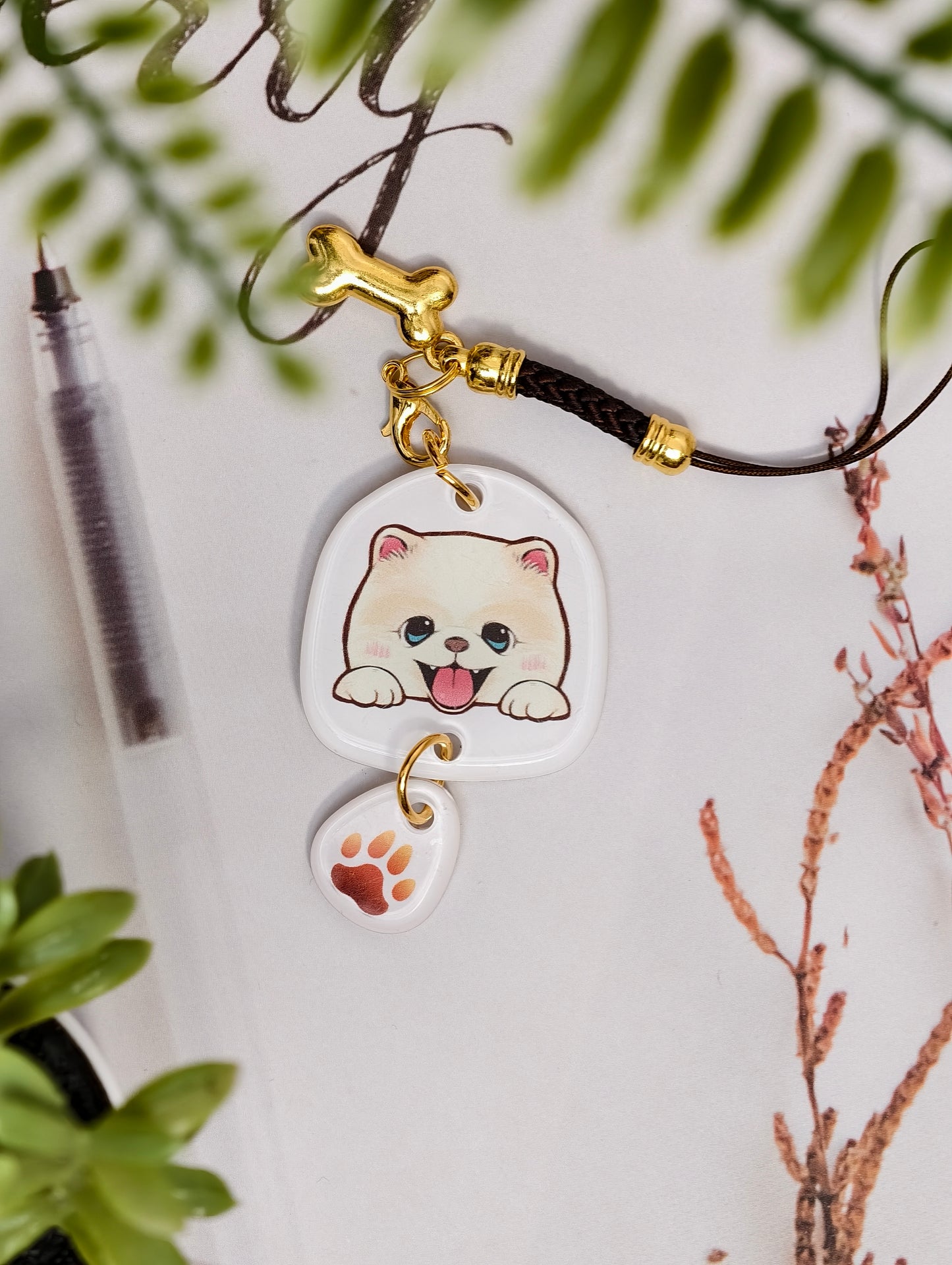 Pomeranian dog illustrated acrylic phone charm with a dangling paw print charm on a light surface with stationery.