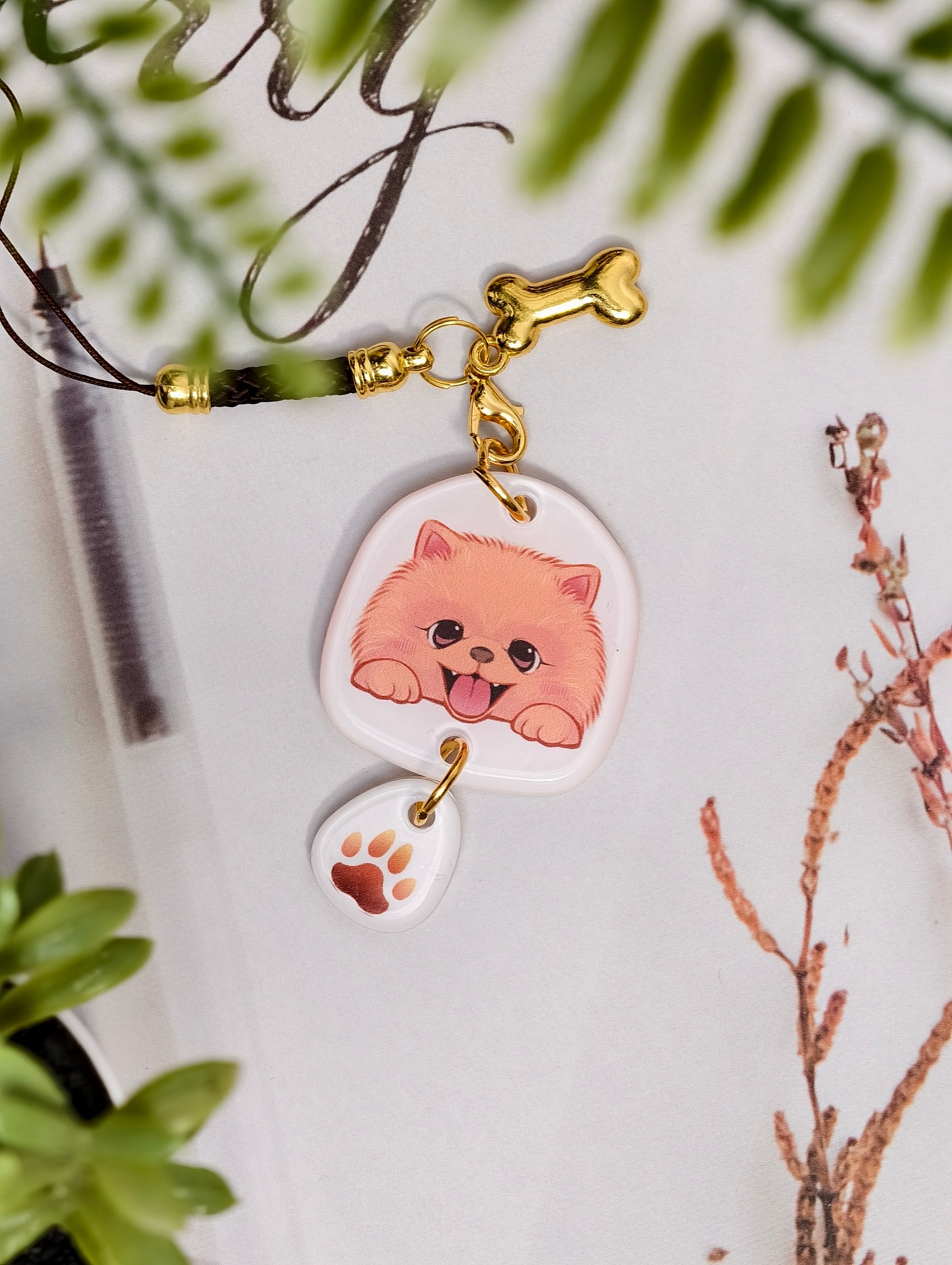 Pomeranian dog illustrated acrylic phone charm with a dangling paw print charm on a light surface with stationery.