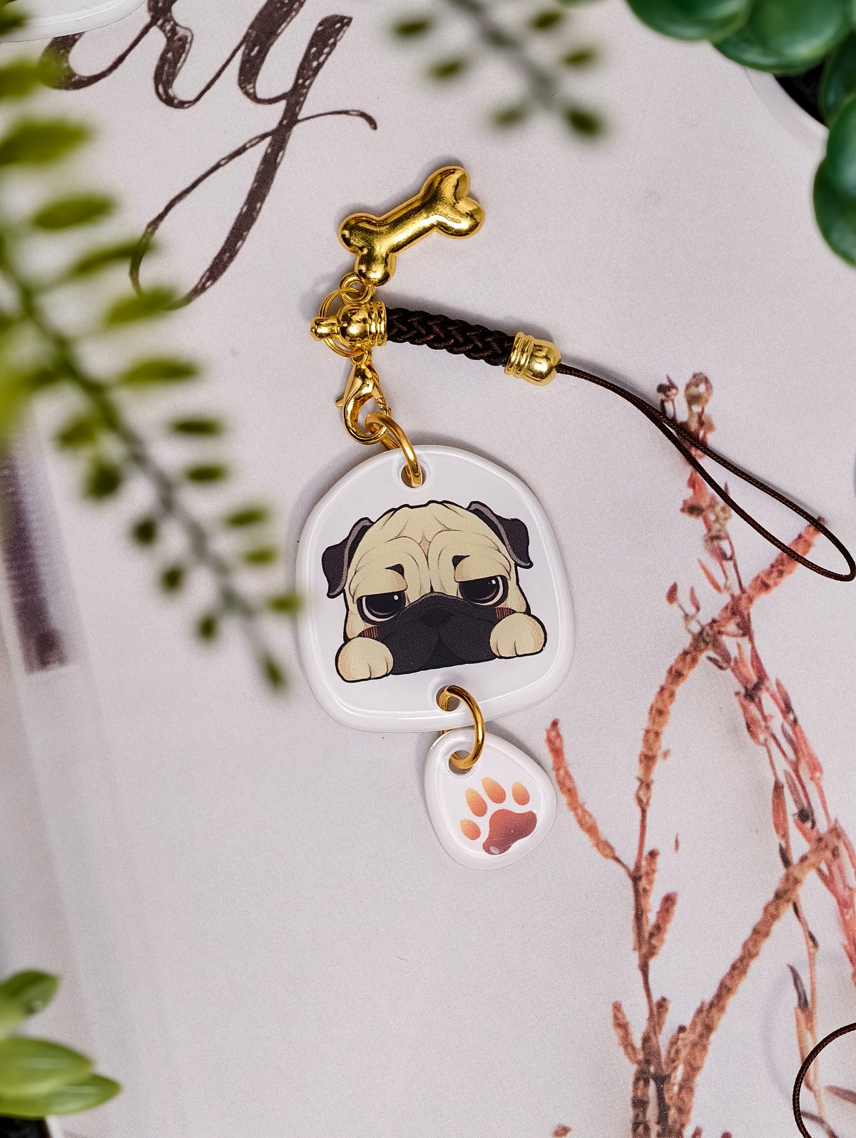 Pug dog illustrated acrylic phone charm with a dangling paw print charm on a light surface with stationery.