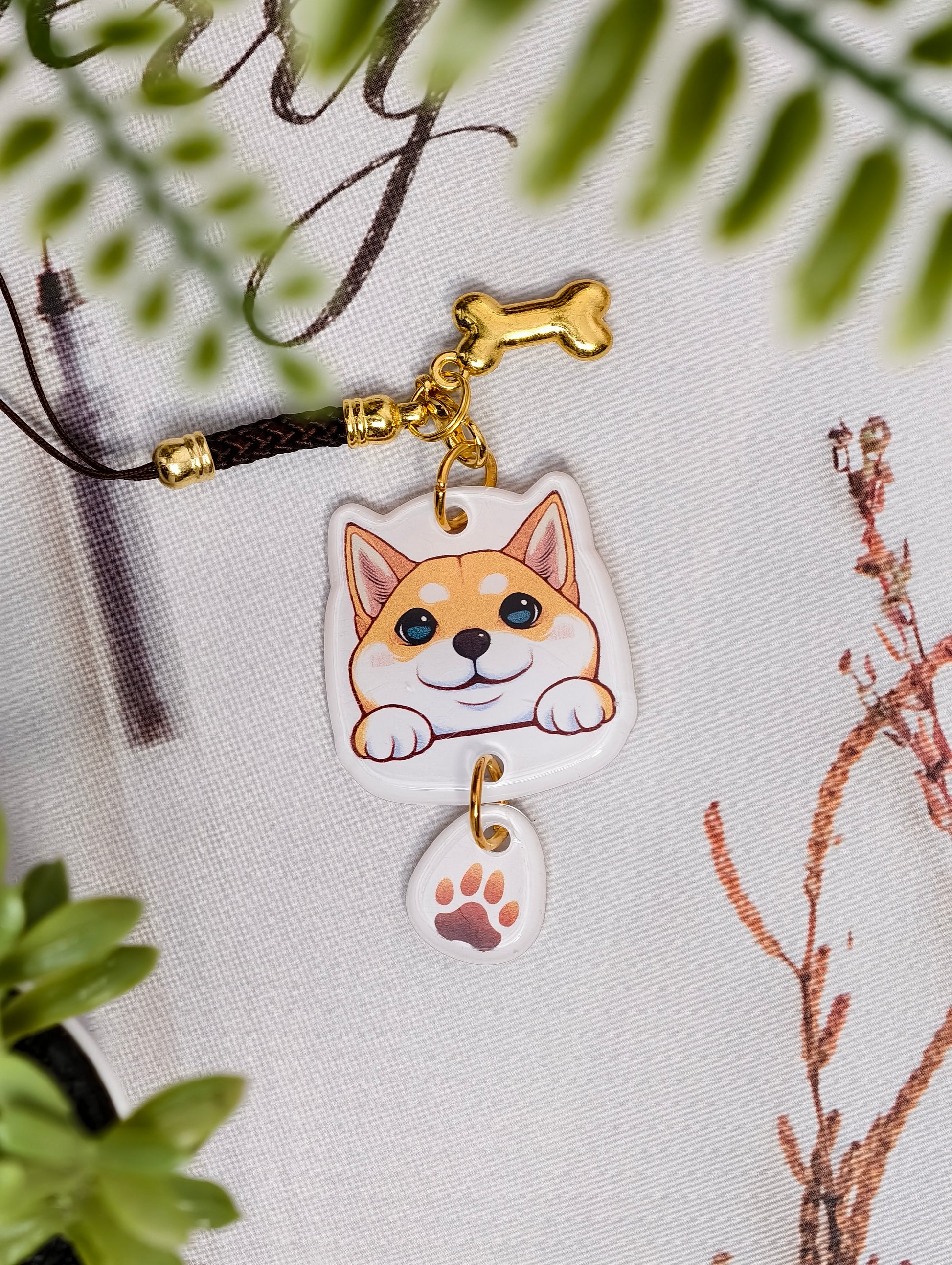 Shiba Inu dog illustrated acrylic phone charm with a dangling paw print charm on a light surface with stationery.